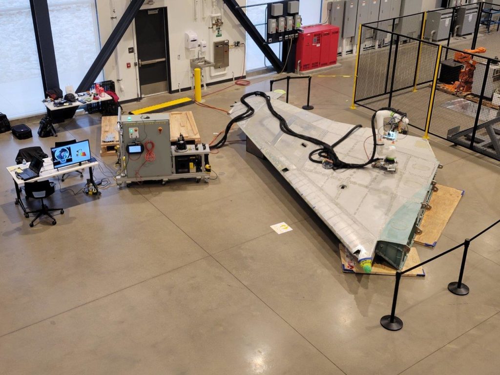 The wing of an F-15 sits on the floor of the Mill 19 high bay with a collaborative robot attached to it
