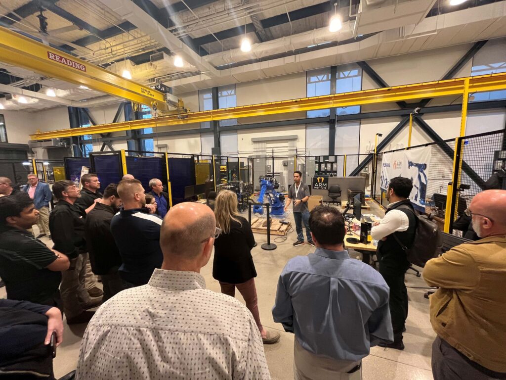 A group of individuals gather in front of a robot as a presenter explains what the industrial robot arm does for manufacturing