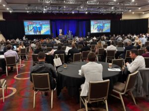 Attendees gather at tables and look at a screen during the Member Meeting