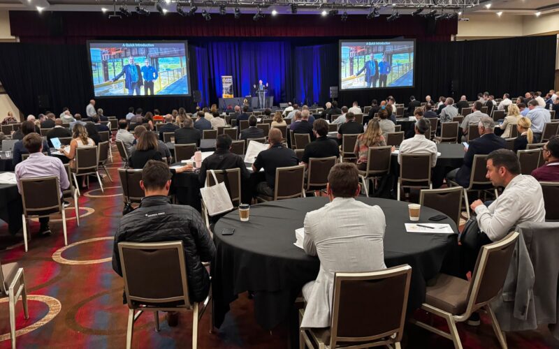 Attendees gather at tables and look at a screen during the Member Meeting